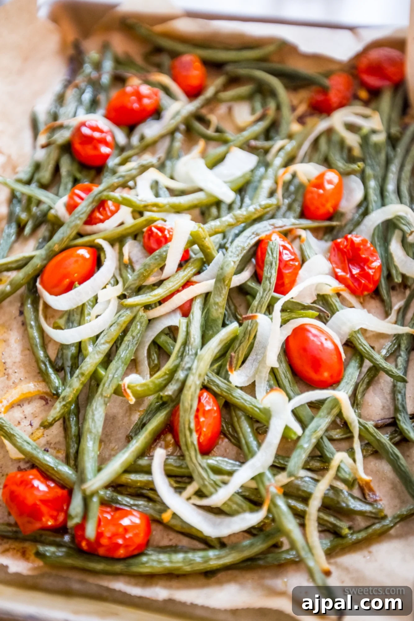 Roasted green beans, tomatoes, and onions glistening on parchment paper in a roasting pan after cooking, ready to be served.