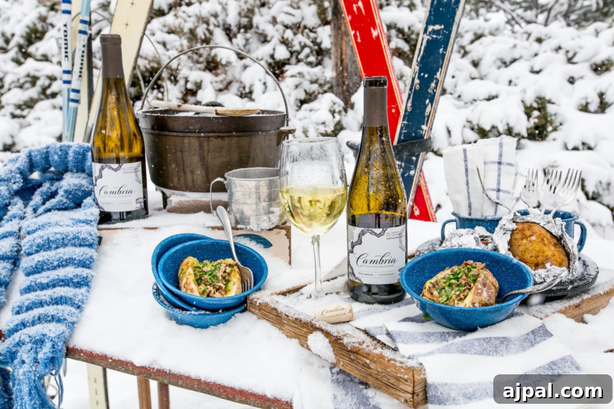 A festive snowy outdoor table setting featuring Cambria wine bottles and bowls of country rib stuffed potatoes, with skis in the background, creating an inviting apres-ski dinner scene.