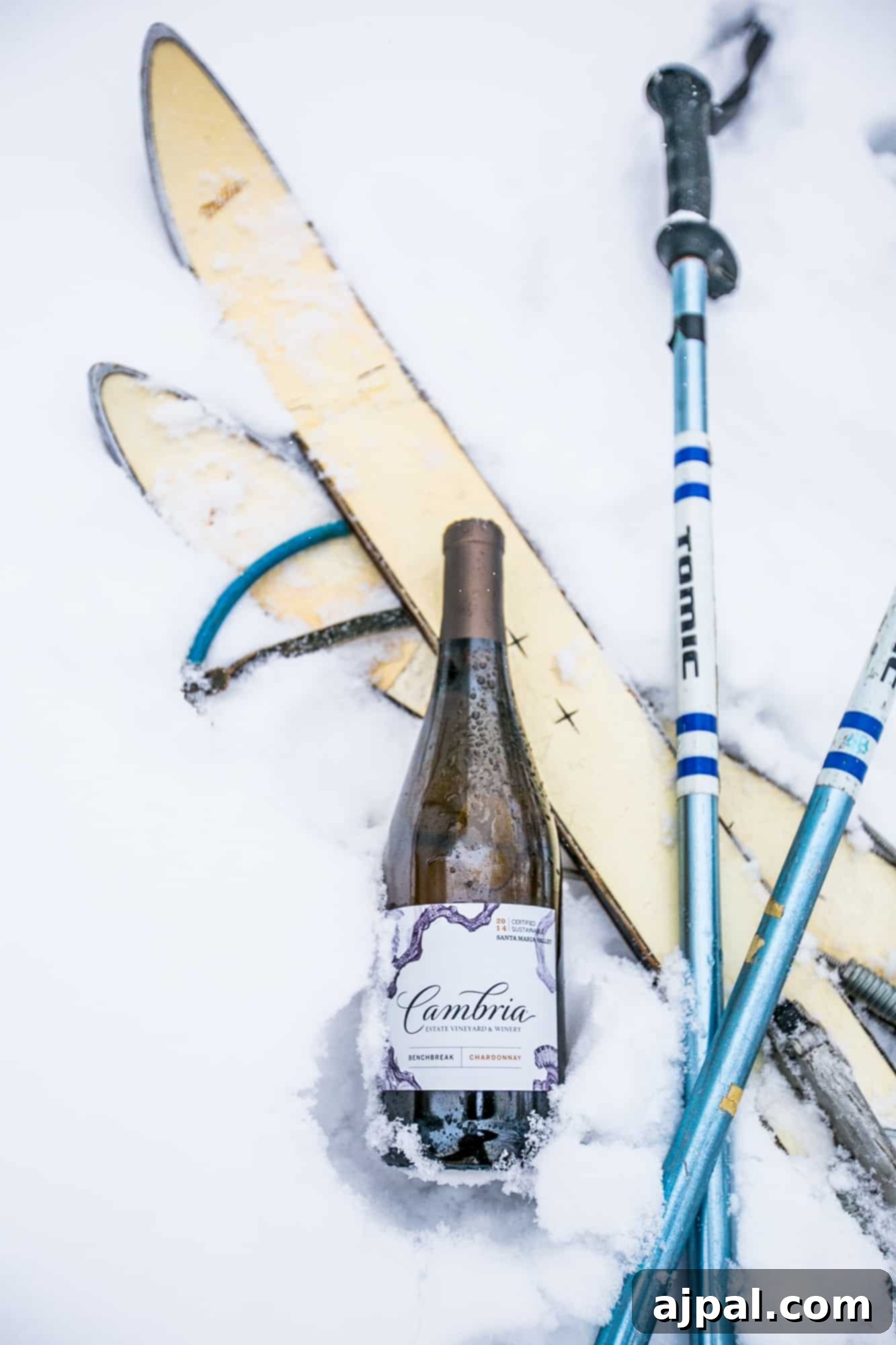 Ski equipment, poles, and a bottle of Cambria wine beautifully arranged in the snow, suggesting a perfect pairing for a ski weekend.