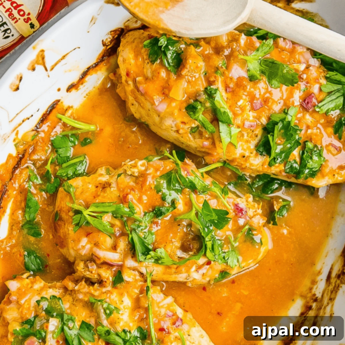 Easy 3 Ingredient Nandos Peri Peri Chicken 5 Three cooked chicken breasts are covered in a 3 Ingredient Nandos Peri Peri Chicken sauce, garnished with chopped parsley in a white baking dish. A wooden spoon rests on the edge of the dish.