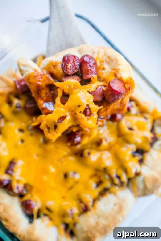 A wooden spoon lifting a serving of Easy Cheesy BBQ Little Smokies Casserole with its golden biscuit base, coated smokies, and melted cheese.