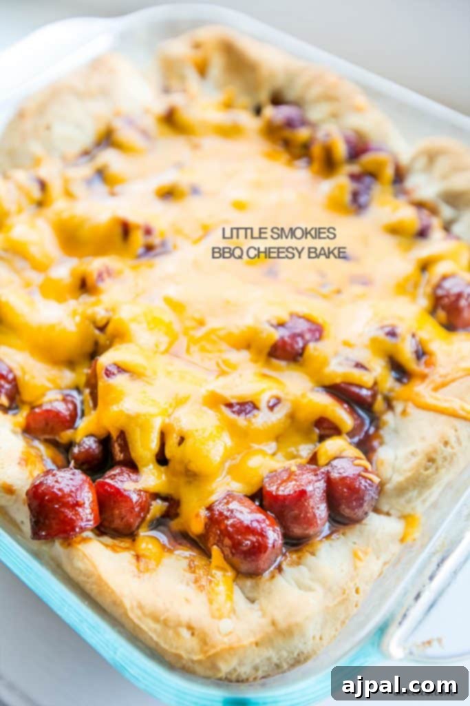 Close-up of golden-brown biscuits topped with BBQ sauce-coated little smokies sausages and bubbly melted cheese in a clear glass baking dish.