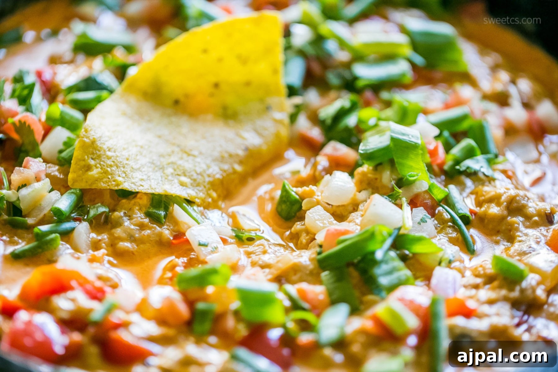 A bowl of rich, creamy chicken enchilada dip garnished with fresh green onions and diced tomatoes, ready for dipping, with tortilla chips on the side.