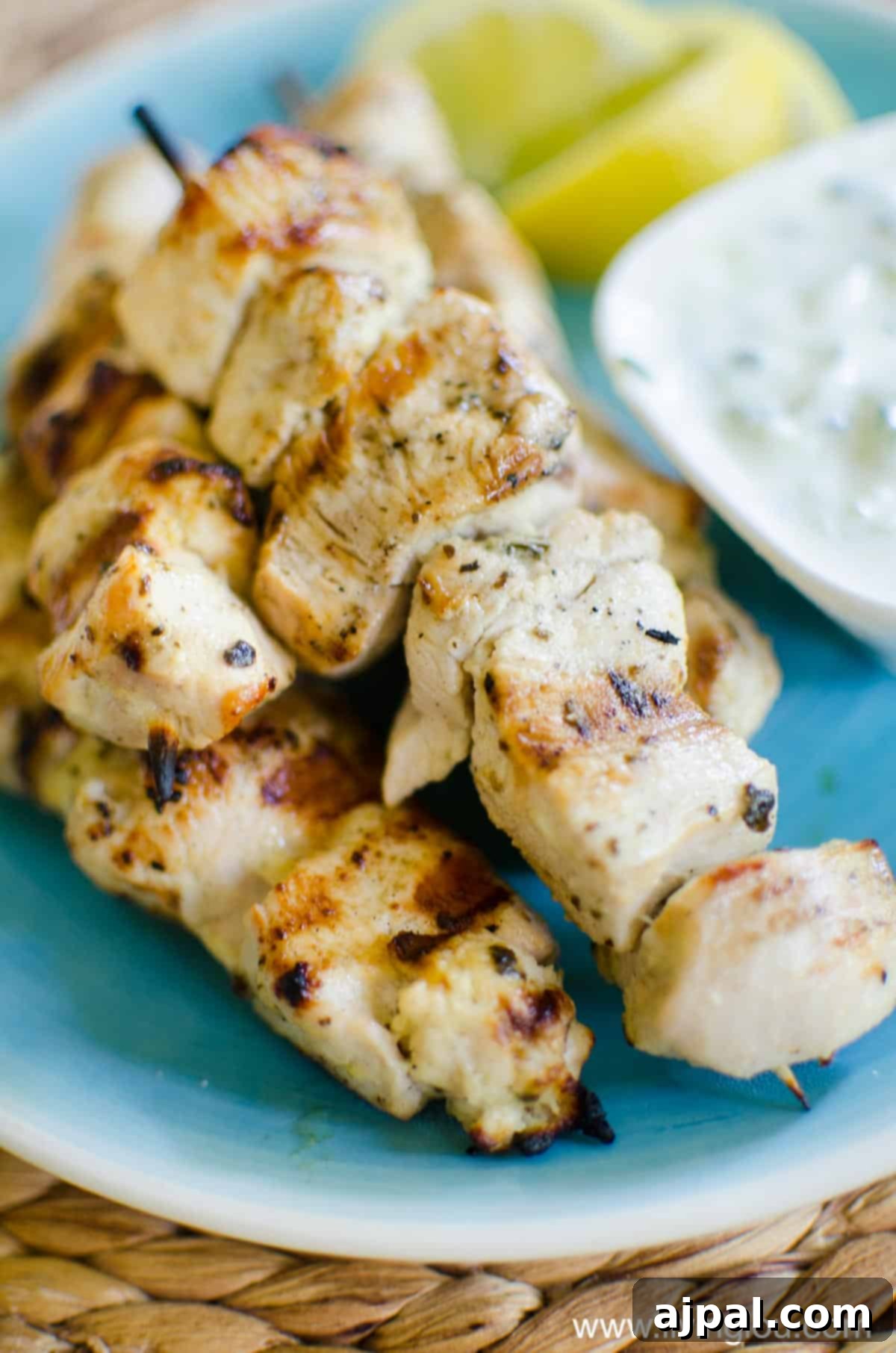 Greek Lemon Herb Chicken Skewers 3 Perfectly grilled Greek chicken souvlaki skewers, garnished with fresh herbs and a lemon wedge, served with a side of creamy tzatziki sauce on a rustic blue plate.