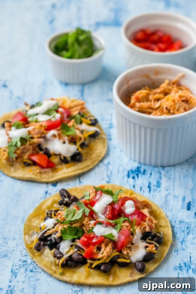 An array of shredded chicken tacos, ready to be customized, with separate bowls of fresh cheese, cilantro, and diced tomatoes on the side.