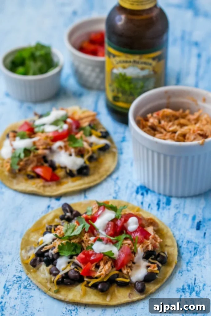 Close-up view of a selection of toppings for shredded chicken tacos, including bowls of grated cheese, fresh cilantro, and diced tomatoes, alongside assembled tacos.