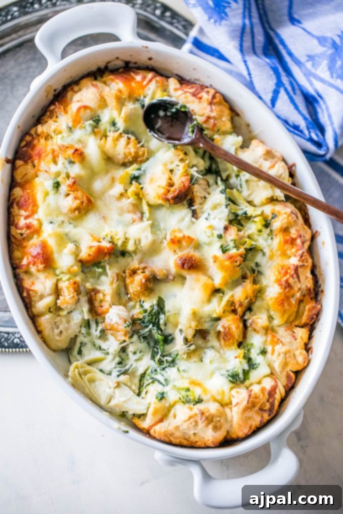A picture of a cheesy chicken, spinach, and artichoke bread dip in a white casserole dish with a wooden spoon in it, ready to be served.