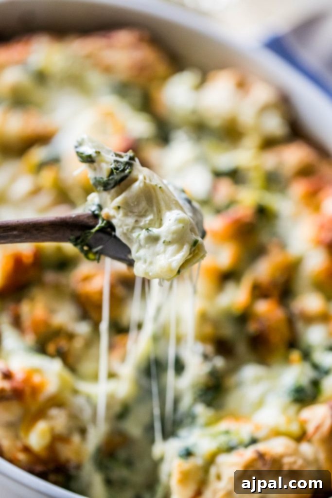 A picture of the cheesy chicken, spinach, and artichoke bread dip in a white casserole dish with a wooden spoon scooping up a portion of the dip.