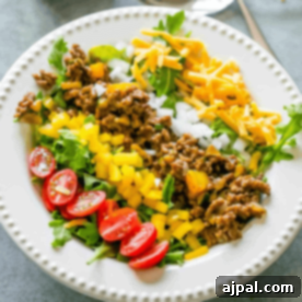 A plate with seasoned ground beef, shredded cheddar cheese, chopped onions, yellow bell peppers, cherry tomatoes, and leafy greens arranged in sections for a fresh low carb taco salad.