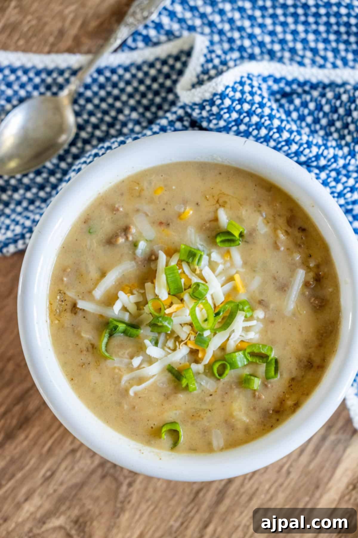 A bowl of creamy hash brown hamburger soup topped with shredded cheese and sliced green onions, placed on a wooden surface with a blue and white patterned cloth and a silver spoon nearby.