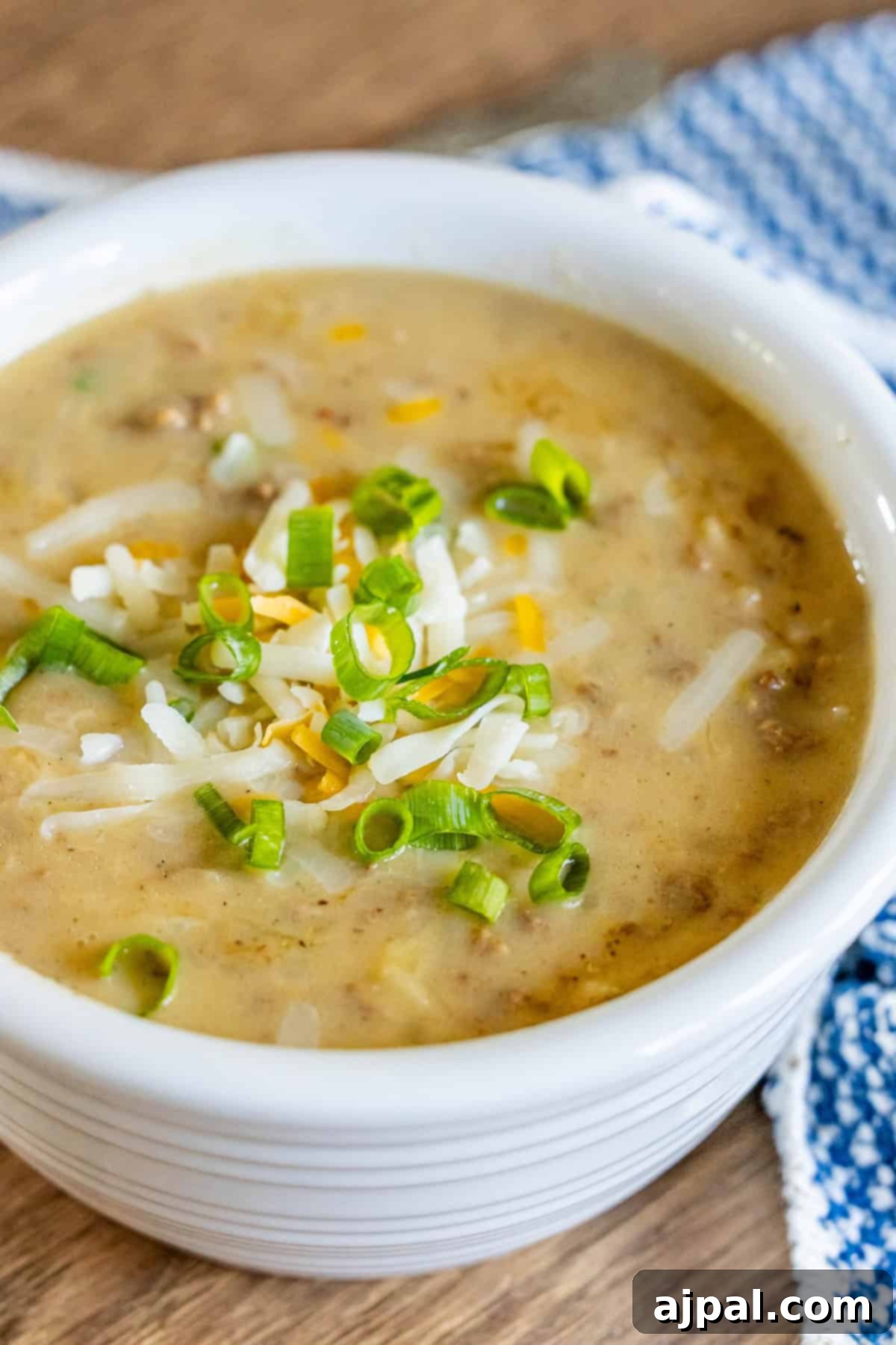 A bowl of creamy hash brown hamburger soup topped with shredded cheese and chopped green onions sits on a wooden table, next to a blue and white checkered cloth.