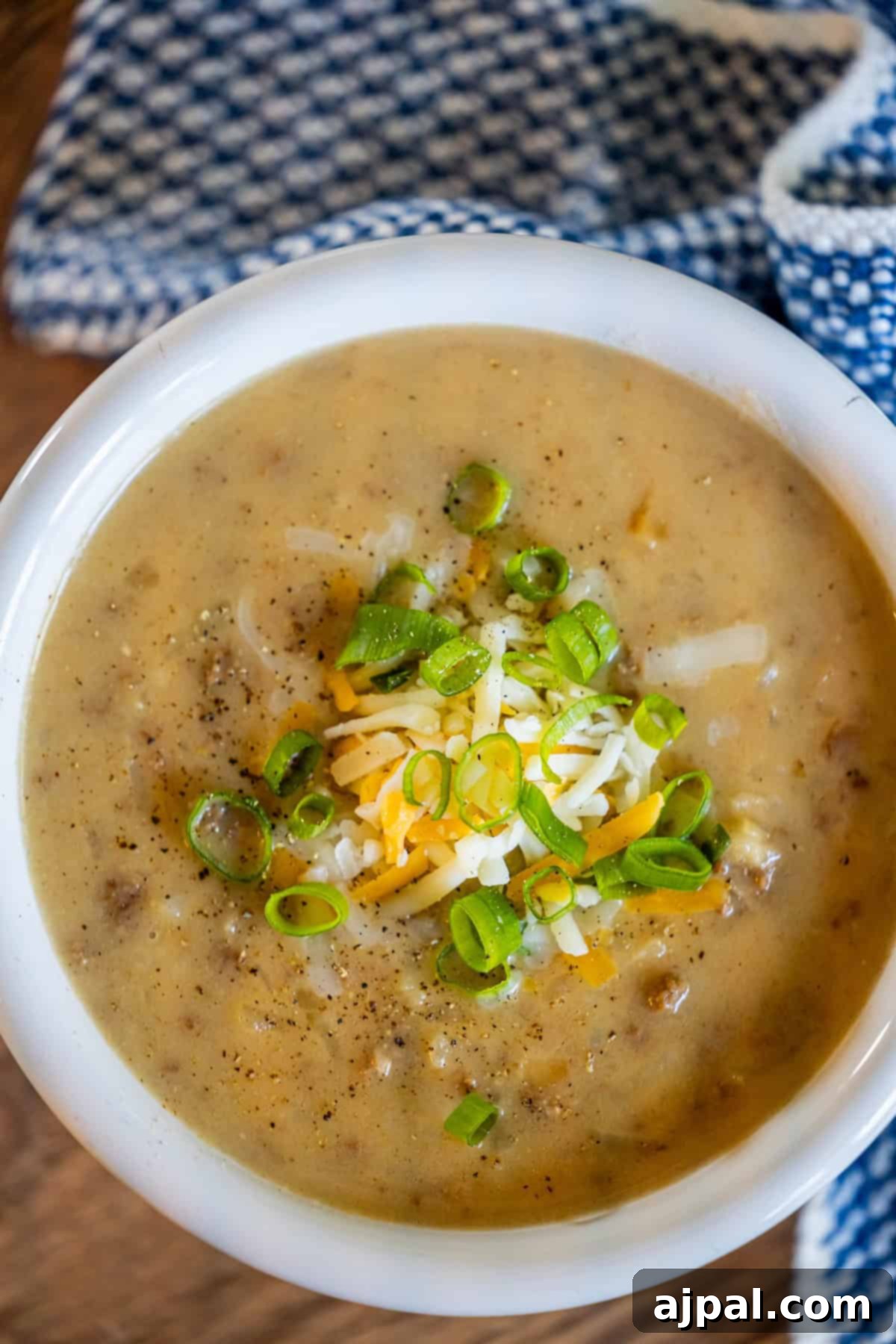 A bowl of creamy hash brown hamburger soup topped with shredded cheese and chopped green onions sits invitingly on a wooden surface, enhanced by the backdrop of a blue-patterned cloth.