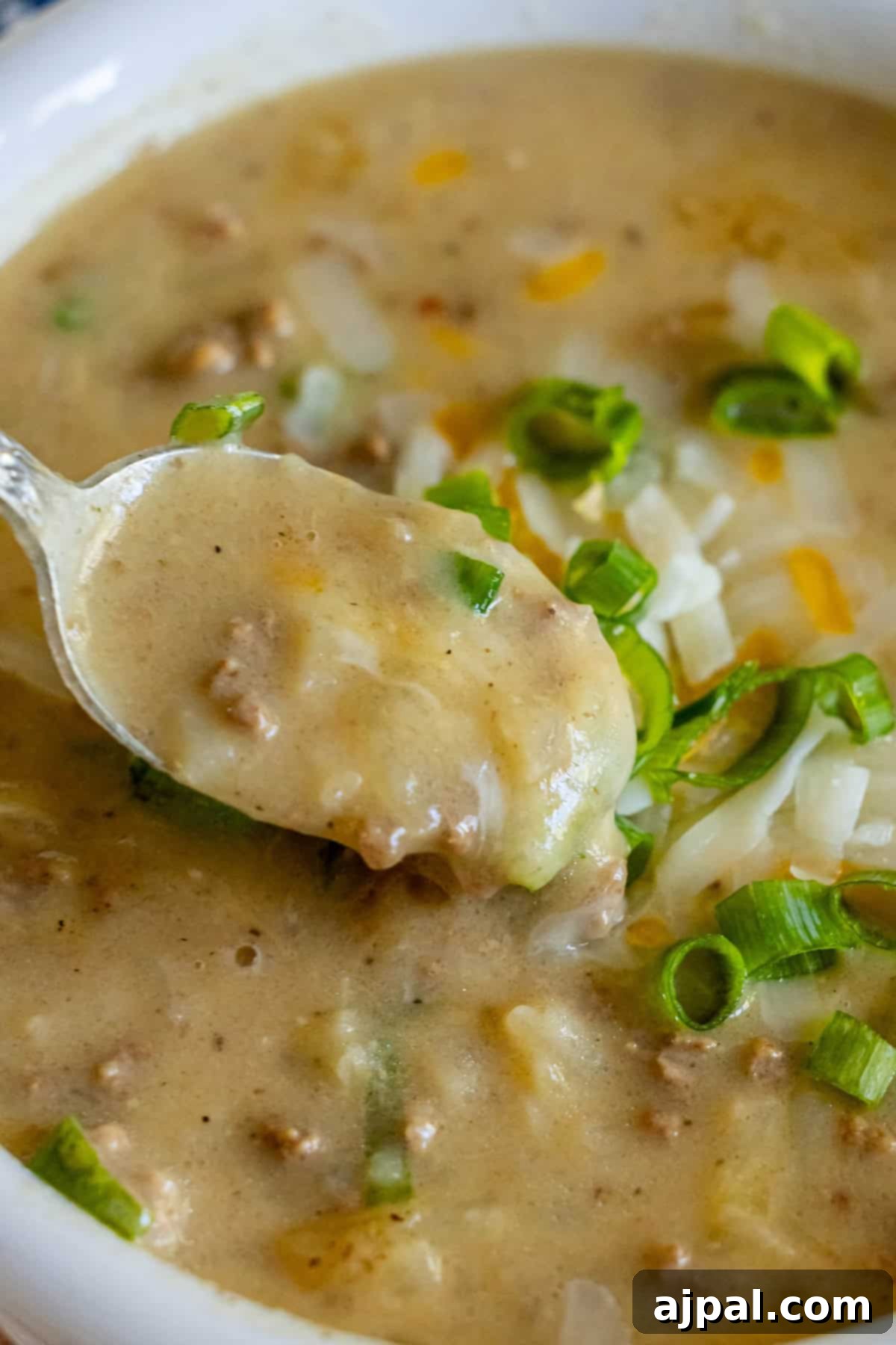 A close-up of creamy Hash Brown Hamburger Soup reveals chunky meat bits, topped with sliced green onions and shredded cheese. A spoon hovers above, poised with a luscious scoop of savory delight.