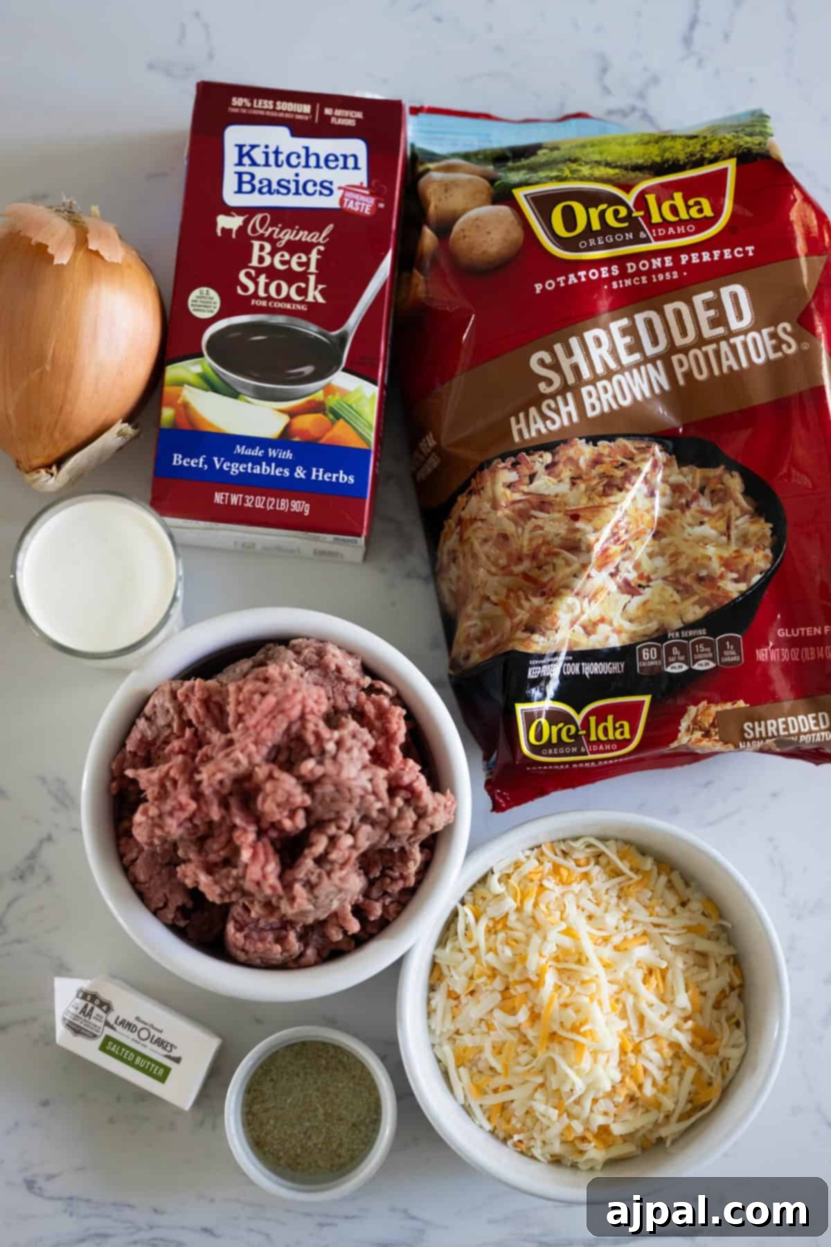 Ingredients on a countertop for a creamy hash brown hamburger soup include onion, beef stock, shredded hash brown potatoes, ground beef, shredded cheese, stick of butter, and a small cup of cream.