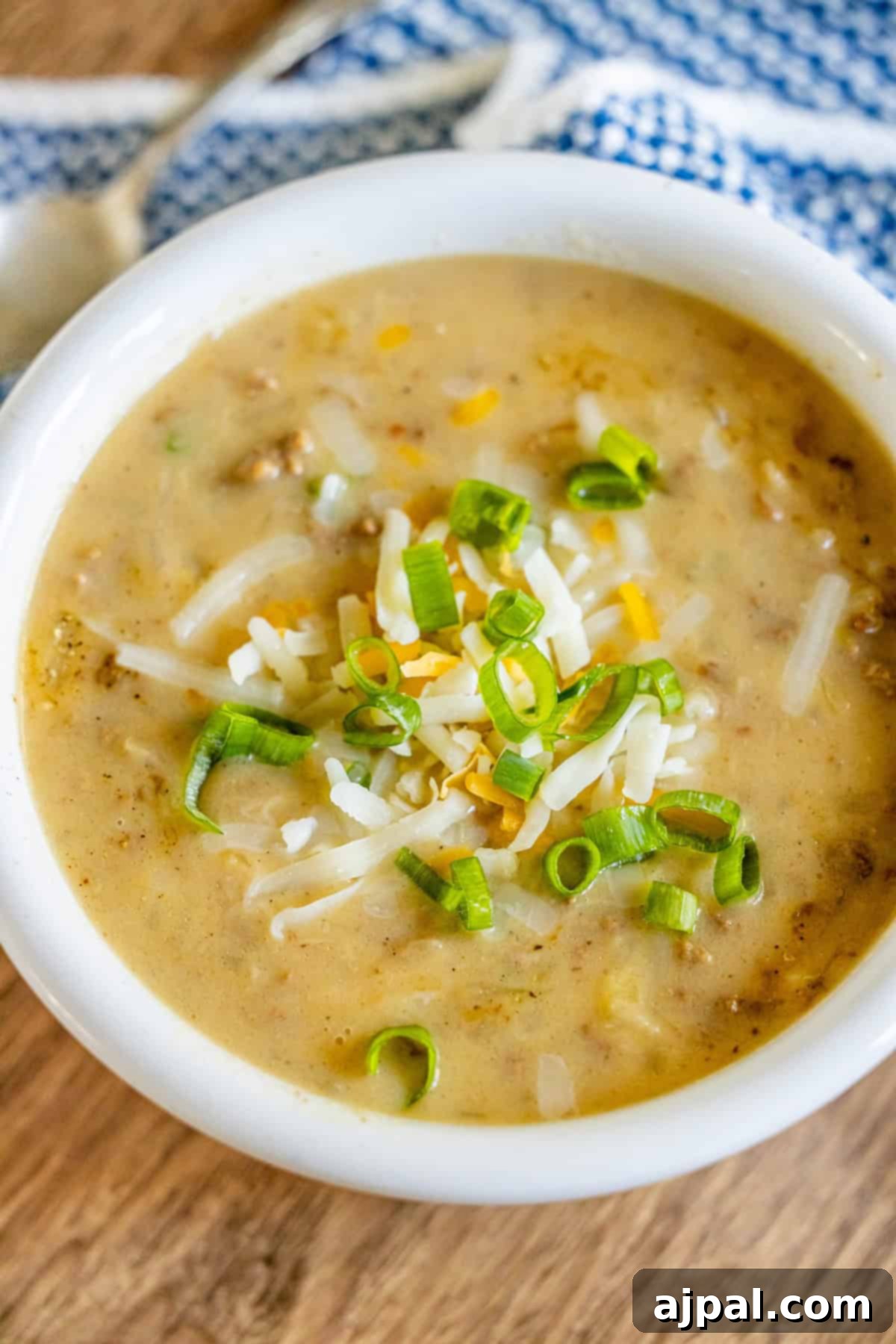 A bowl of creamy hash brown hamburger soup topped with shredded cheese and chopped green onions rests on a wooden surface, accompanied by a blue and white cloth and a spoon nearby.