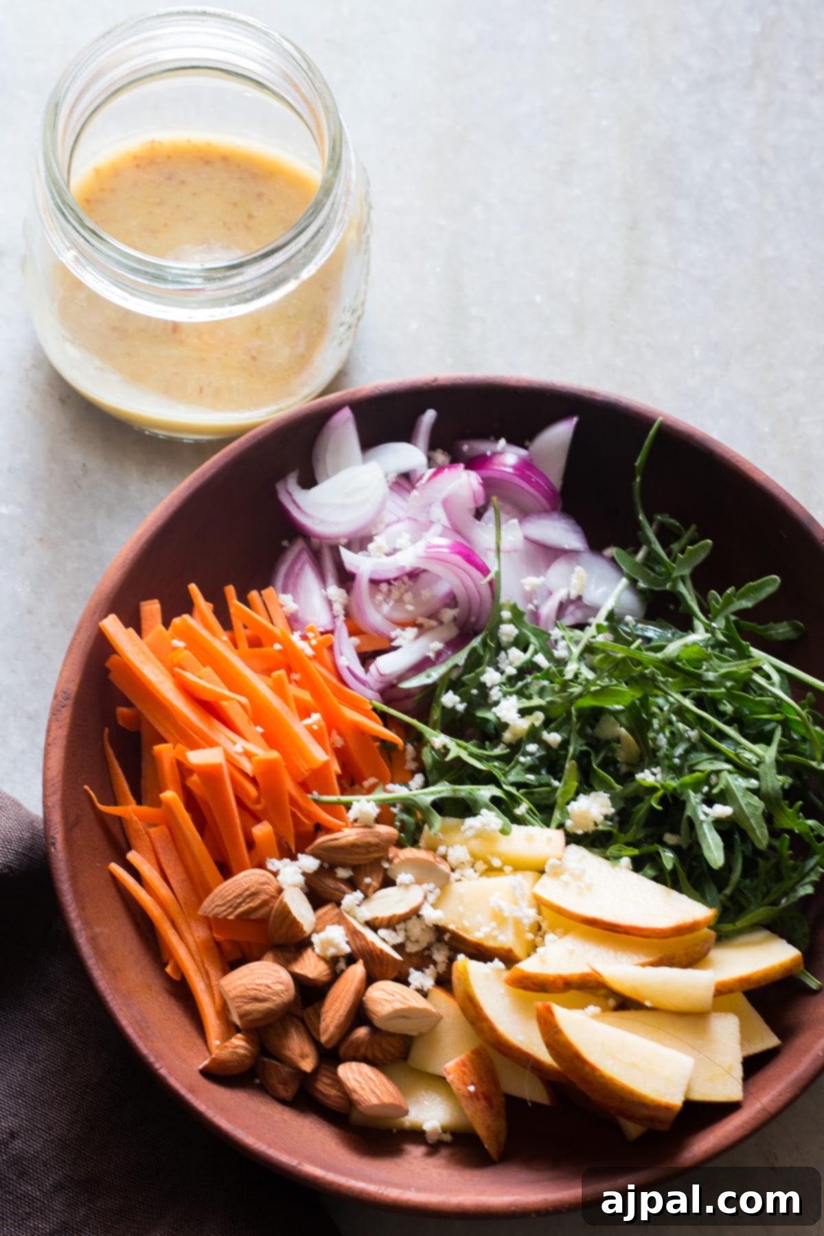 Orange Kissed Apple Arugula Almond Salad 2 A rustic wooden bowl filled with vibrant apple slices, crunchy almonds, fresh carrots, peppery arugula, thinly sliced red onion, and creamy goat cheese. This healthy and delicious fall salad is topped with a bright orange dressing.