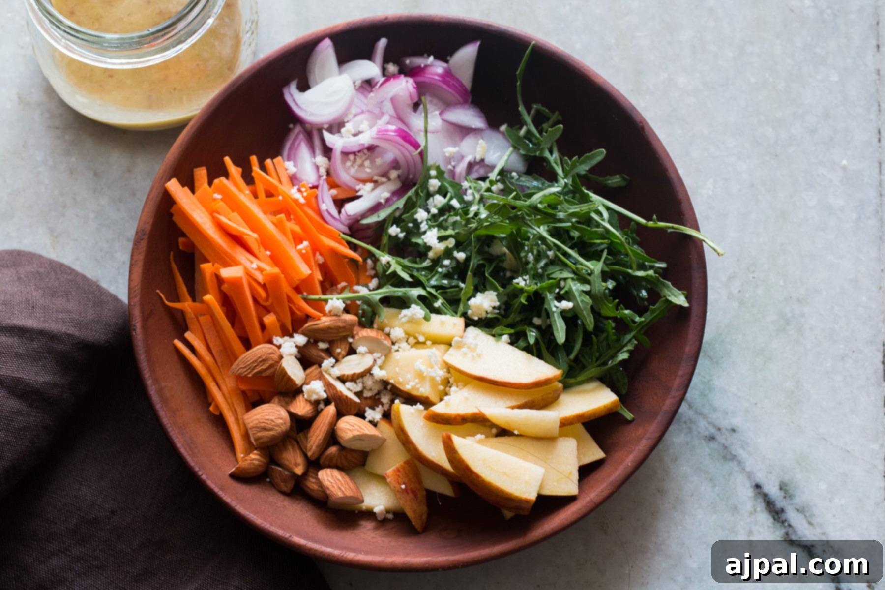 Orange Kissed Apple Arugula Almond Salad 5 A rustic wooden bowl filled with vibrant apple slices, crunchy almonds, fresh carrots, peppery arugula, thinly sliced red onion, and creamy goat cheese.