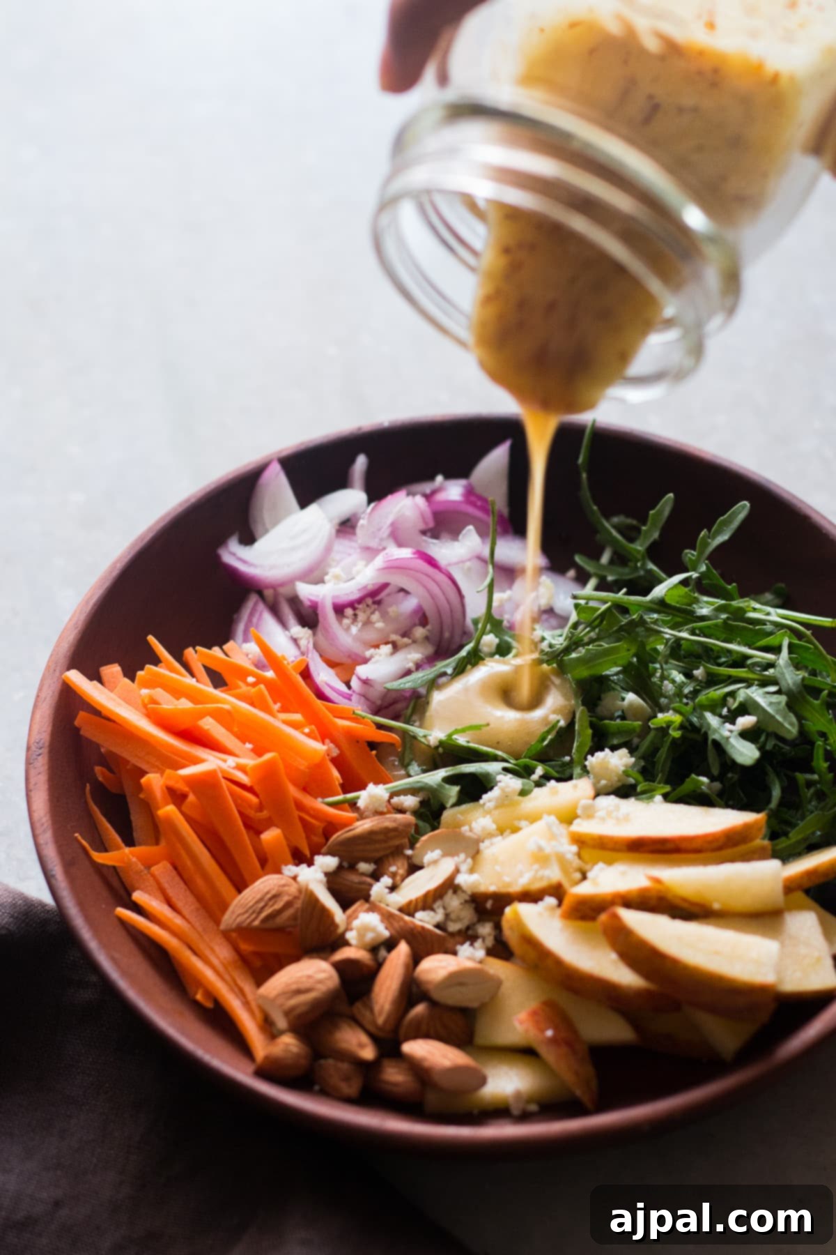 Orange Kissed Apple Arugula Almond Salad 3 A wooden bowl holds a vibrant mix of apple slices, almonds, carrots, arugula, and goat cheese, with a creamy orange dressing being generously poured over the fresh ingredients.