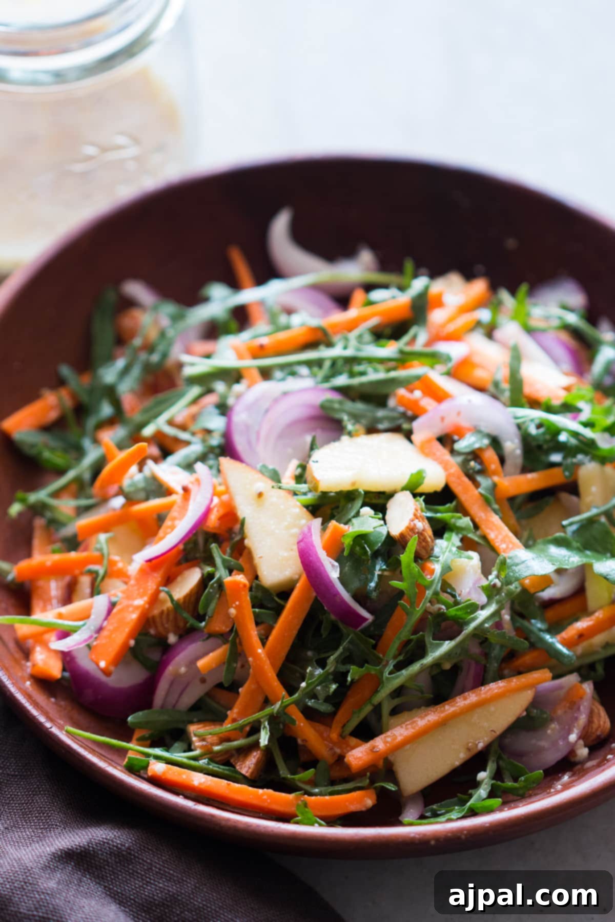 Orange Kissed Apple Arugula Almond Salad 4 A rustic wooden bowl holds a thoroughly mixed salad featuring vibrant apple slices, crunchy almonds, fresh carrots, peppery arugula, thinly sliced red onion, and creamy goat cheese, all coated in a rich orange dressing.