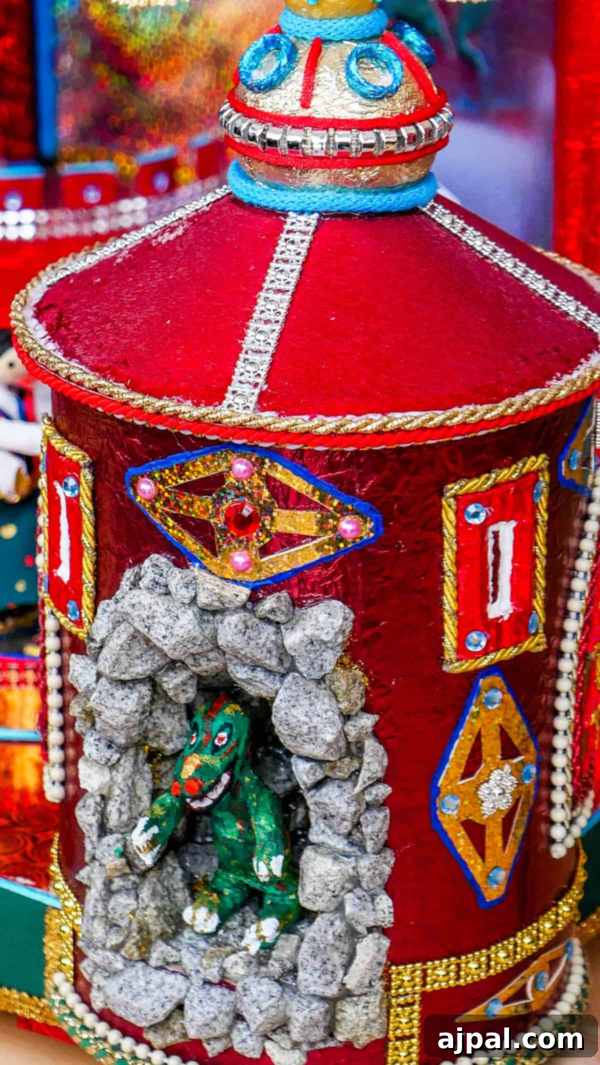 This colorful decorative object, reminiscent of entries in the Krakow Nativity Competition, features a red cylindrical structure adorned with intricate patterns and jewels. A small green dragon figurine sits nestled into a stone-like cavity on the side, adding an element of mythical charm.