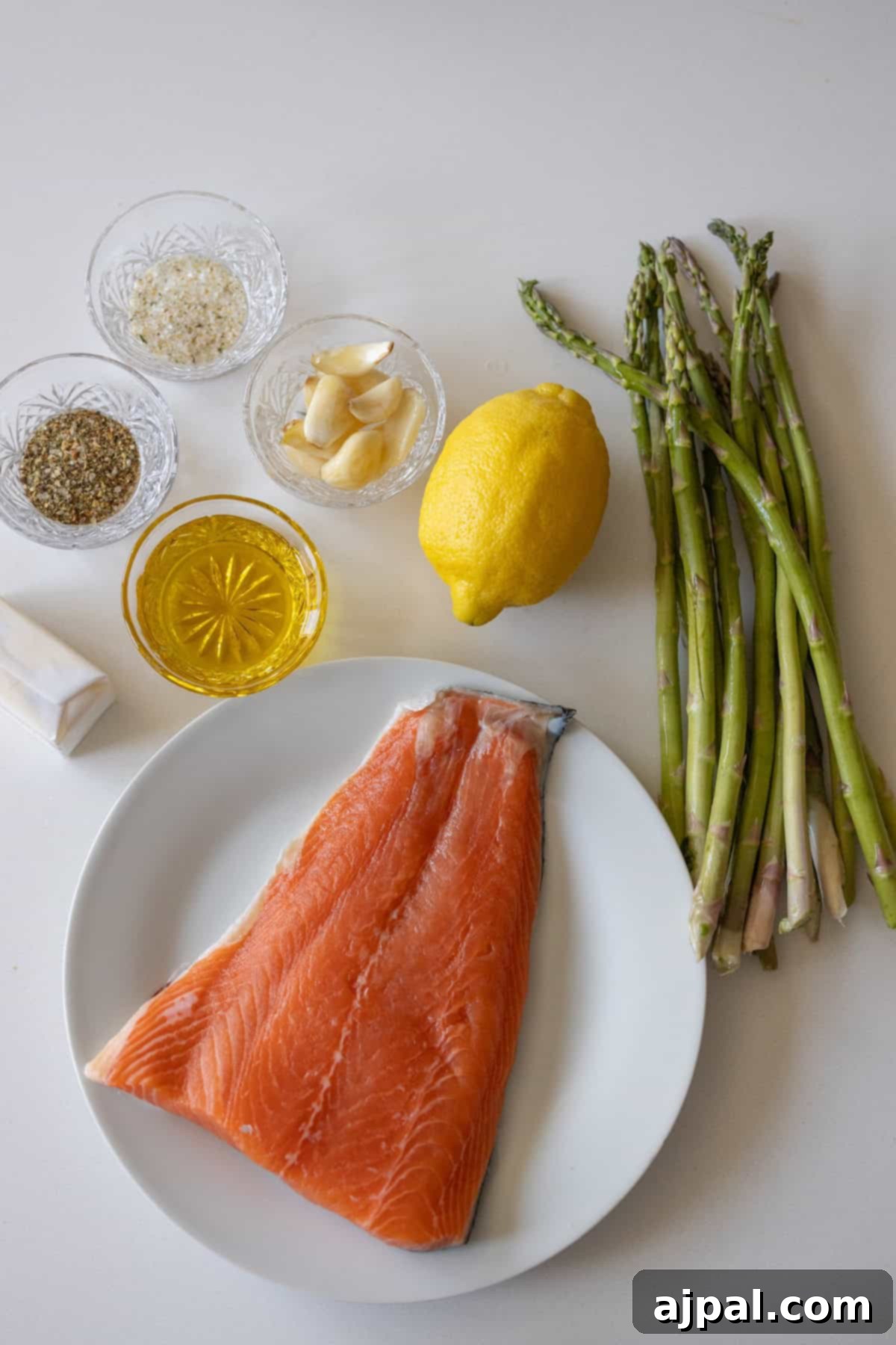 Zesty Sheet Pan Lemon Garlic Salmon and Asparagus 3 A raw salmon fillet on a white plate is surrounded by fresh asparagus, lemon, garlic cloves, olive oil, butter, salt, and spices, all ready for an easy salmon and asparagus recipe.