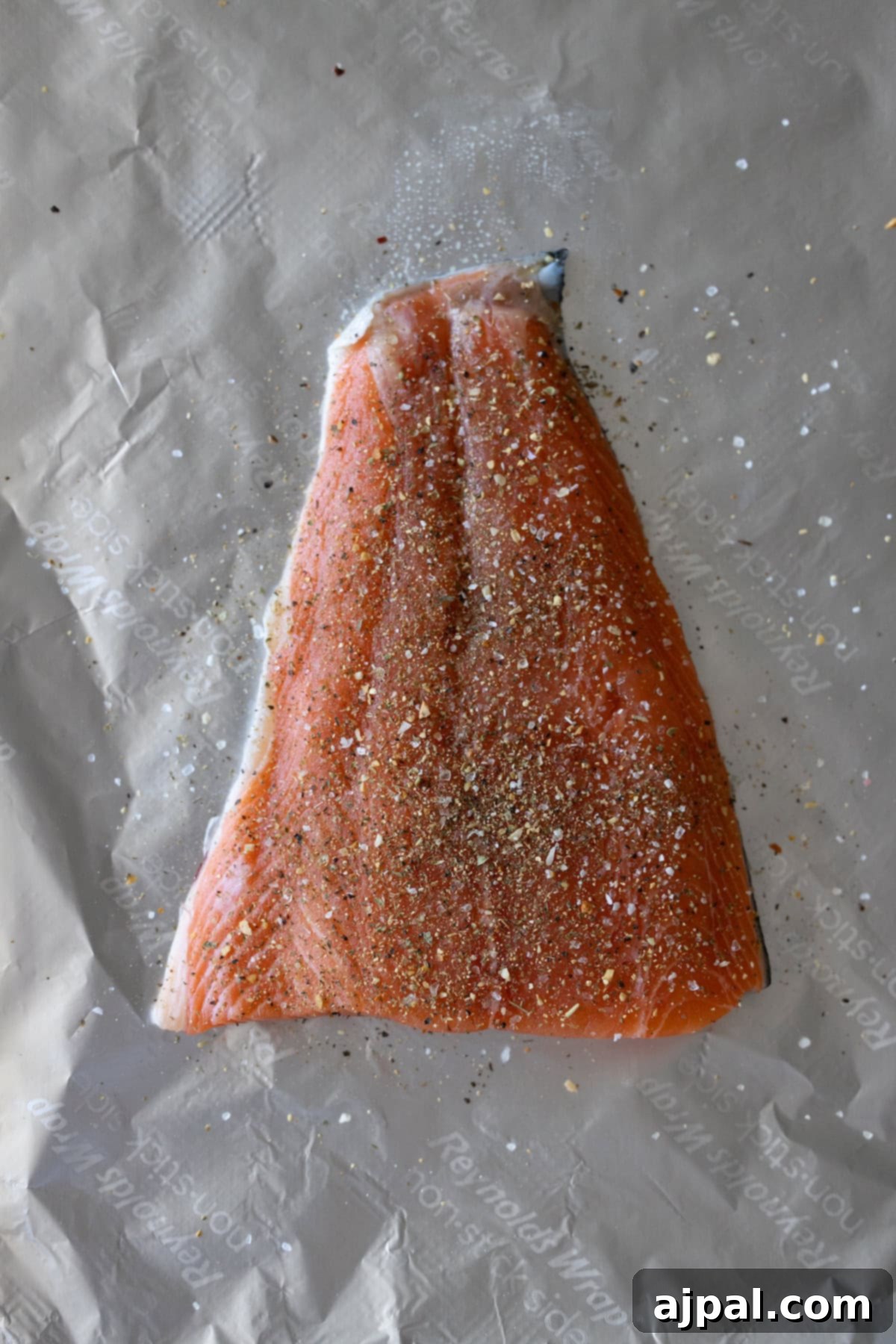 Zesty Sheet Pan Lemon Garlic Salmon and Asparagus 4 A raw salmon fillet generously sprinkled with ground black pepper and other seasonings, resting on parchment paper, ready to be cooked as part of a delicious Sheet Pan Lemon Garlic Salmon and Asparagus dinner.