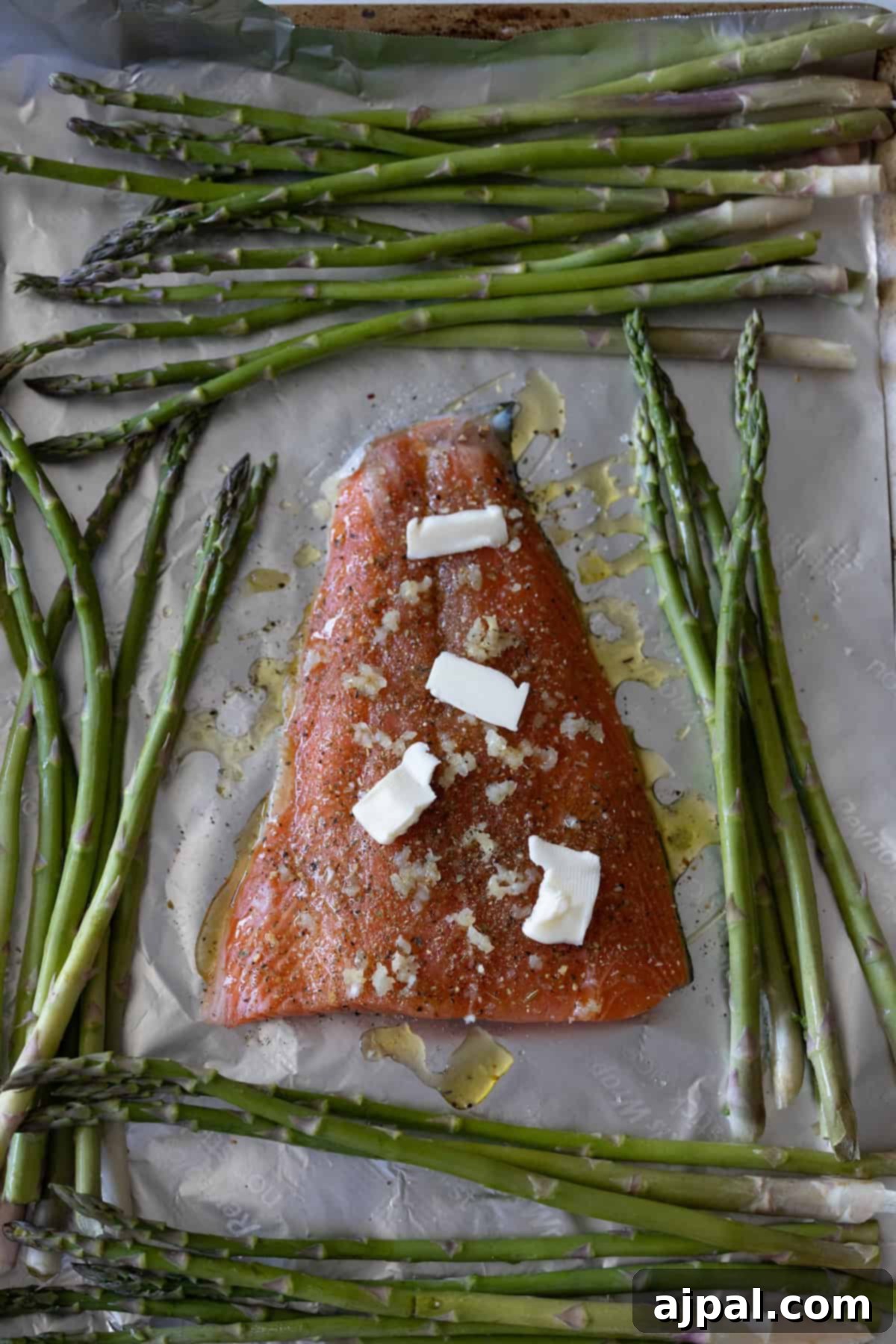 Zesty Sheet Pan Lemon Garlic Salmon and Asparagus 6 A raw salmon fillet topped with butter and minced garlic rests on parchment, surrounded by fresh asparagus spears, creating a perfect setup for an easy salmon and asparagus recipe.