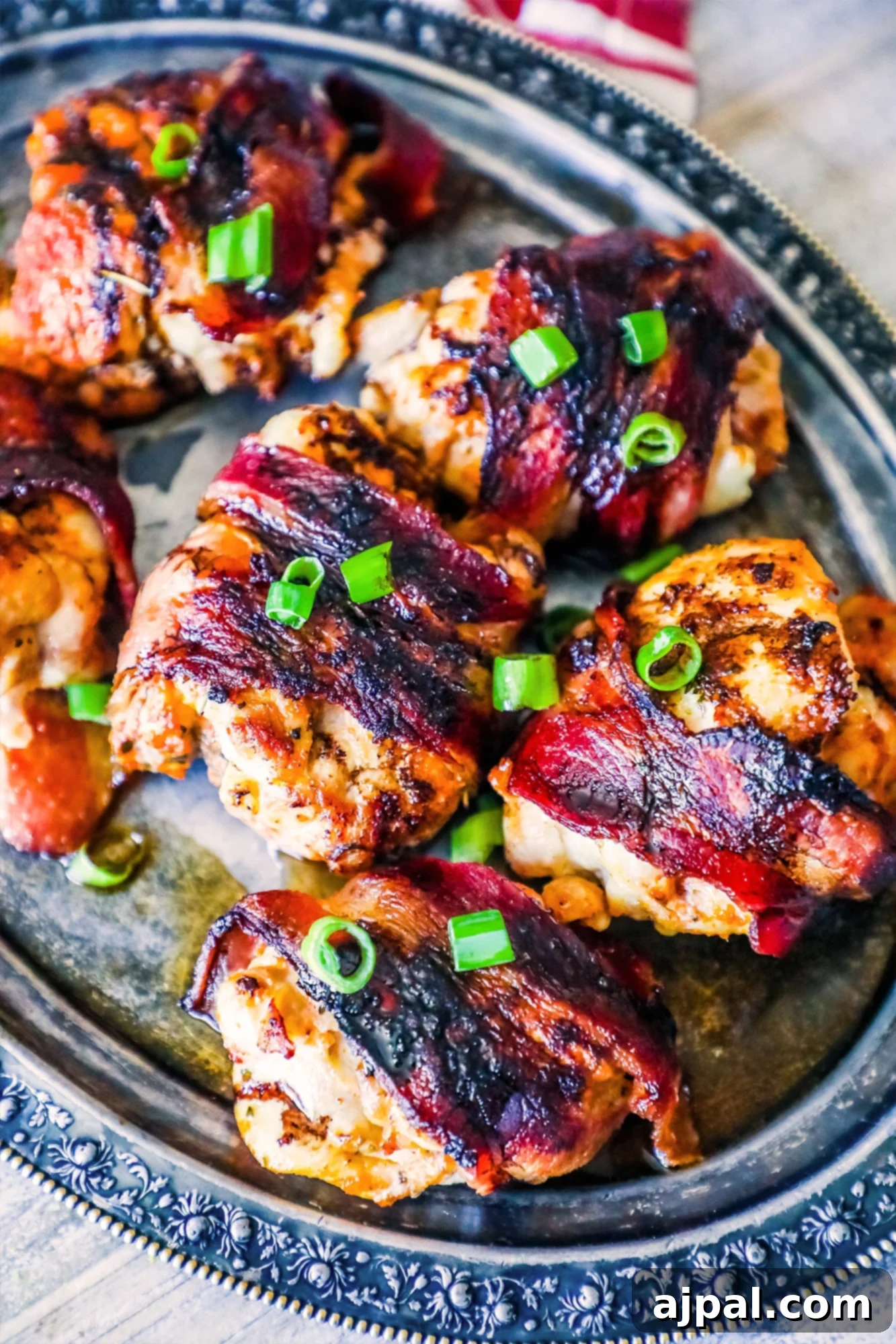 Sweet and Savory Balsamic Bacon Chicken Thighs 3 Bacon-wrapped chicken thighs beautifully arranged on a silver serving plate, accented with vibrant green onions.