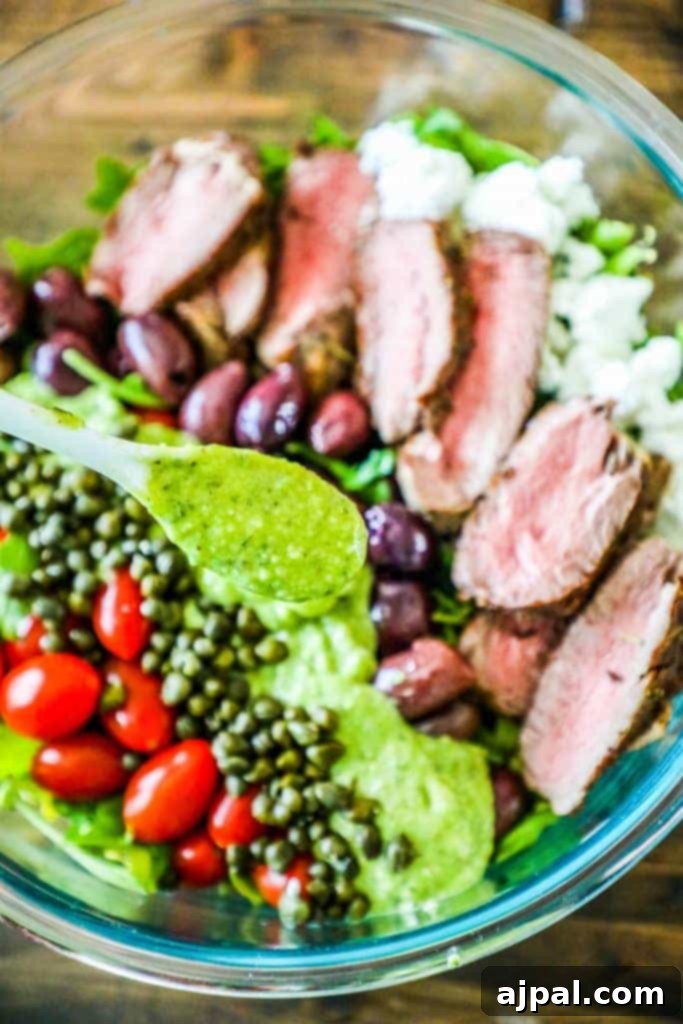 A beautifully arranged bowl of creamy avocado steak salad, featuring tender slices of steak, fresh lettuce, juicy cherry tomatoes, briny capers, and ripe olives, all drizzled with a rich green avocado dressing.