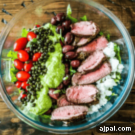 A bowl filled with sliced beef, cherry tomatoes, kalamata olives, lentils, cauliflower rice, and a dollop of creamy avocado sauce.
