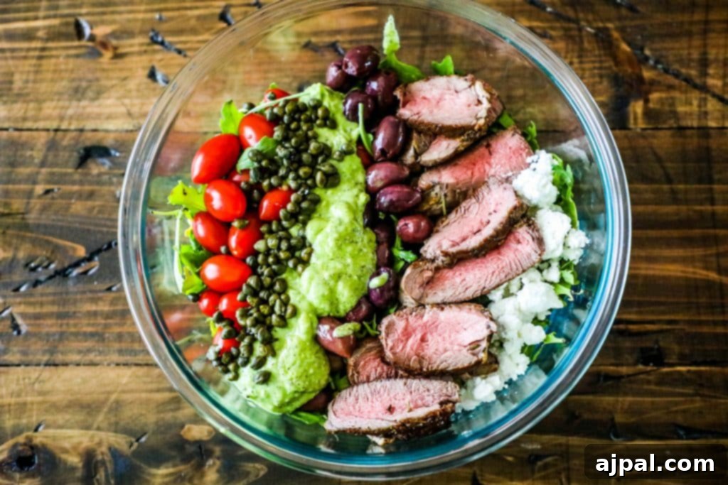 Close-up shot of creamy avocado steak salad, showcasing the rich green dressing and the vibrant mix of steak, capers, tomatoes, olives, and feta cheese.