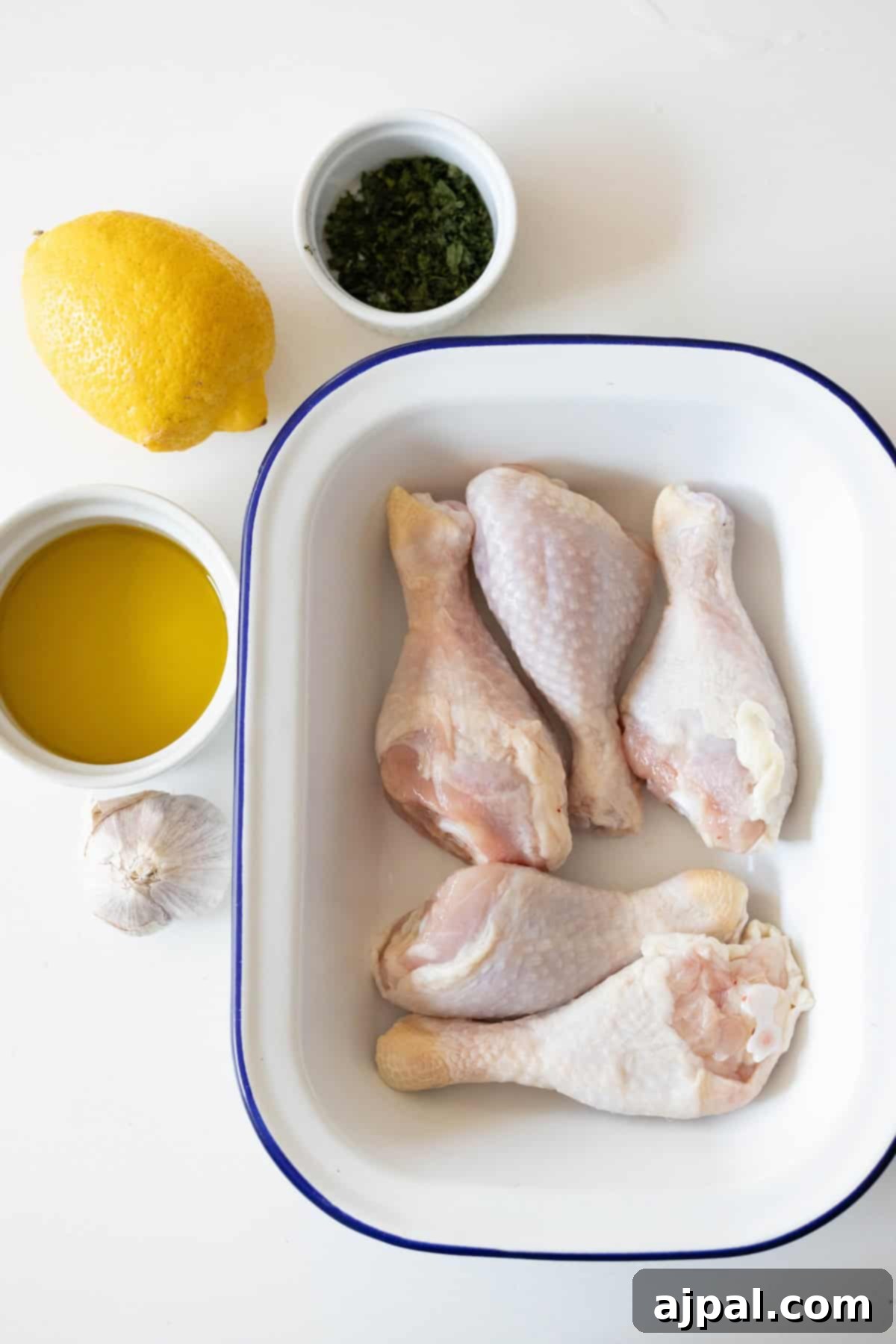 Zesty Greek Chicken Drumsticks 6 Greek Chicken Drumsticks nestle in a white dish, surrounded by a lemon, a bowl of olive oil, garlic, and a small cup of chopped herbs on a pristine white surface.