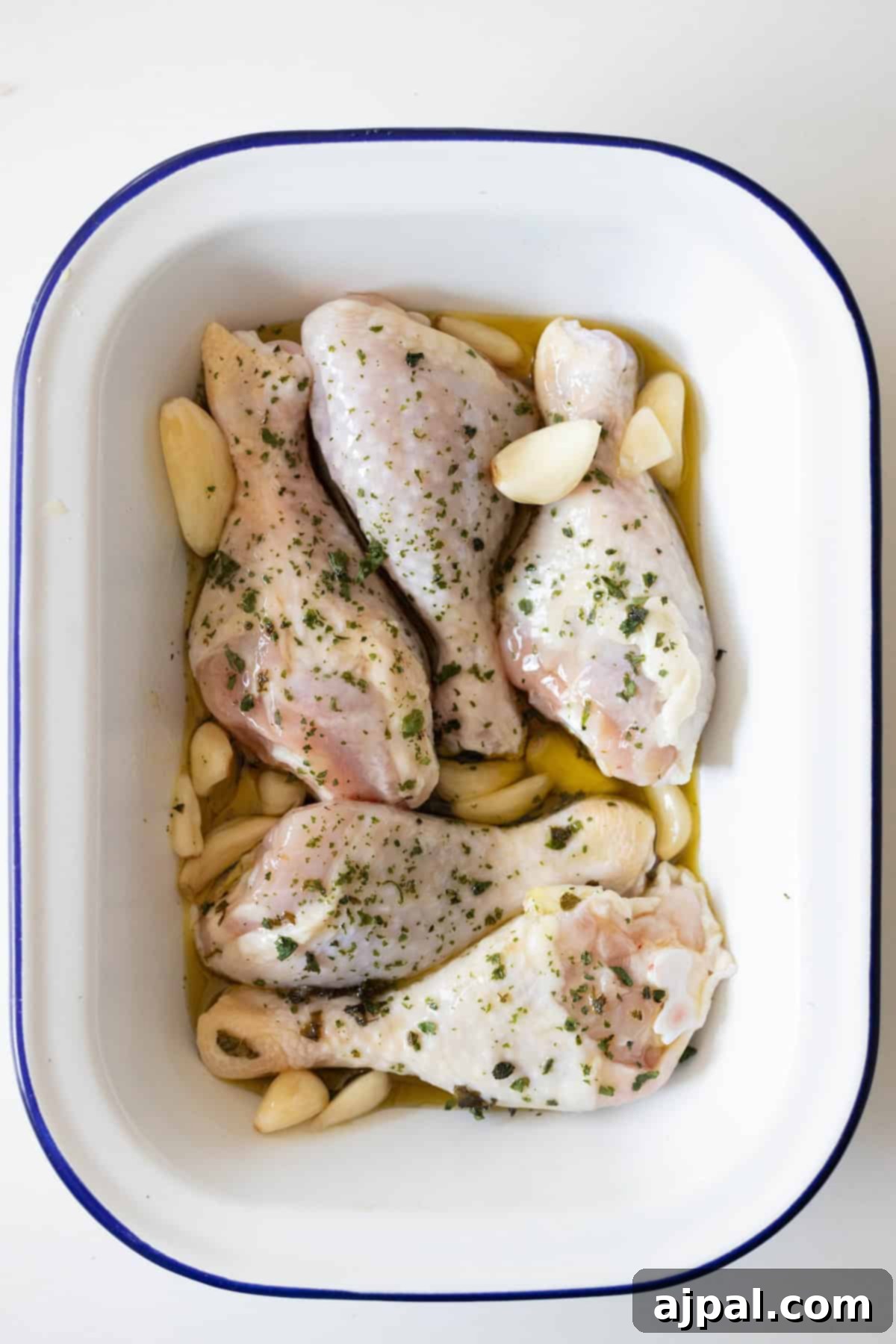 Zesty Greek Chicken Drumsticks 7 Greek chicken drumsticks marinated with herbs and garlic cloves in a white baking dish.