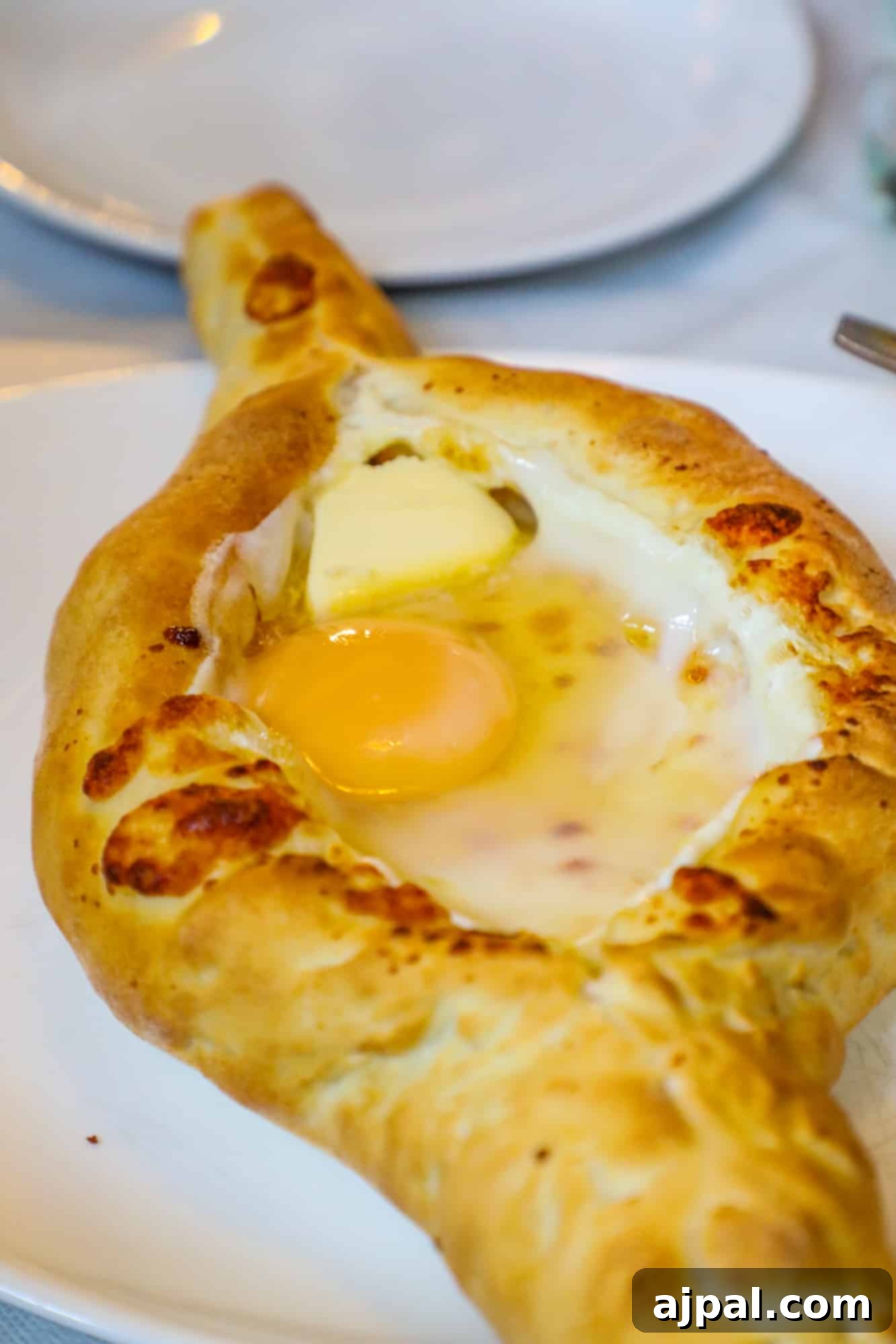 A close-up of Adjaruli Khachapuri, a boat-shaped Georgian cheese bread topped with a soft-cooked egg and butter, served on a white plate.