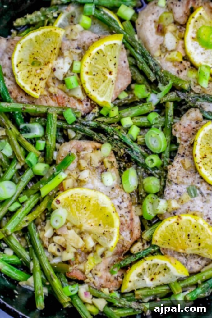 A vibrant close-up of a sizzling one-pot lemon garlic pork chop and asparagus skillet meal. Golden-brown pork chops are nestled amongst bright green asparagus spears and thin lemon slices, all glistening in a rich, buttery sauce. Minced garlic bits are visible, adding to the savory aroma.
