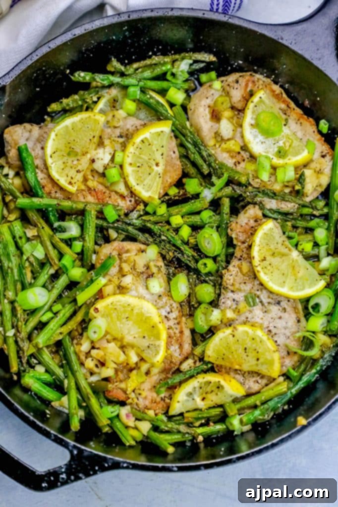 A top-down view of a sizzling one-pot lemon garlic pork chop and asparagus skillet. Golden-brown pork chops are artfully arranged with bright green asparagus spears, fresh lemon wedges, and a rich, aromatic pan sauce. The meal is garnished with fresh herbs, ready for serving.