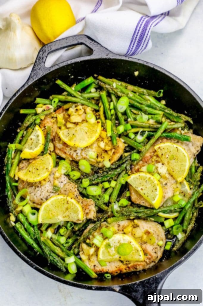 A close-up shot of a sizzling cast-iron skillet filled with perfectly cooked lemon garlic pork chops and asparagus. The pork chops are golden-brown, adorned with thin lemon slices, while the bright green asparagus spears are tender-crisp. A rich, savory pan sauce coats everything, ready to be served.
