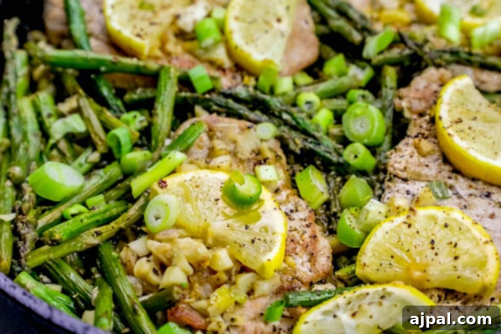 An overhead shot of a one-pot lemon garlic pork chop and asparagus skillet. The golden-brown pork chops are garnished with fresh lemon slices and green herbs, while the vibrant green asparagus spears are perfectly cooked and coated in a light, savory sauce. It's a tempting, wholesome meal.