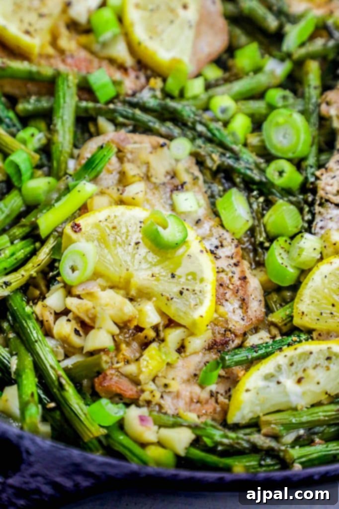 A large cast-iron skillet filled with a delectable one-pan lemon garlic pork chop dinner. Three perfectly seared pork chops are arranged with vibrant green asparagus spears and bright yellow lemon slices, all coated in a rich, savory pan sauce, ready to be served.