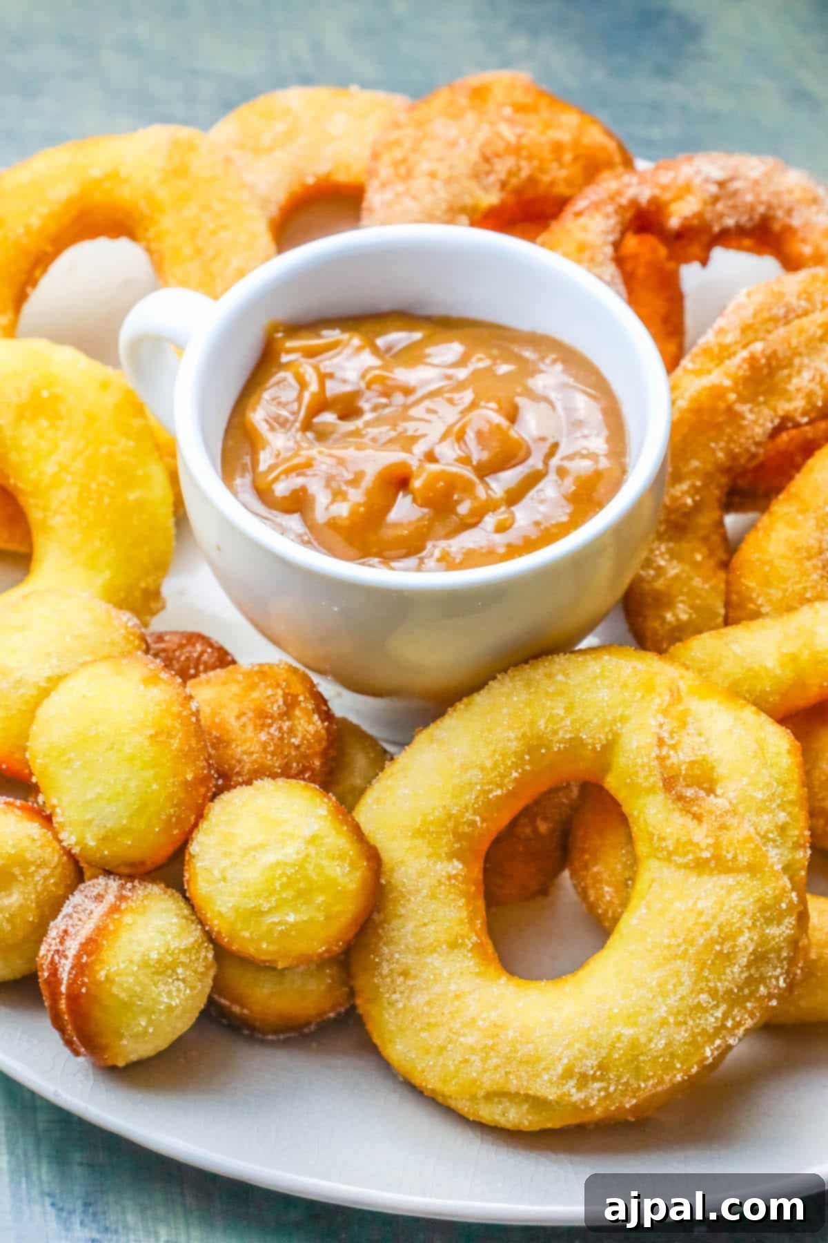 A delightful arrangement of freshly fried 2-ingredient mulling spiced donuts and their smaller donut holes, artfully placed around a small bowl of sweet caramel sauce. The scene evokes a cozy fall breakfast setting.