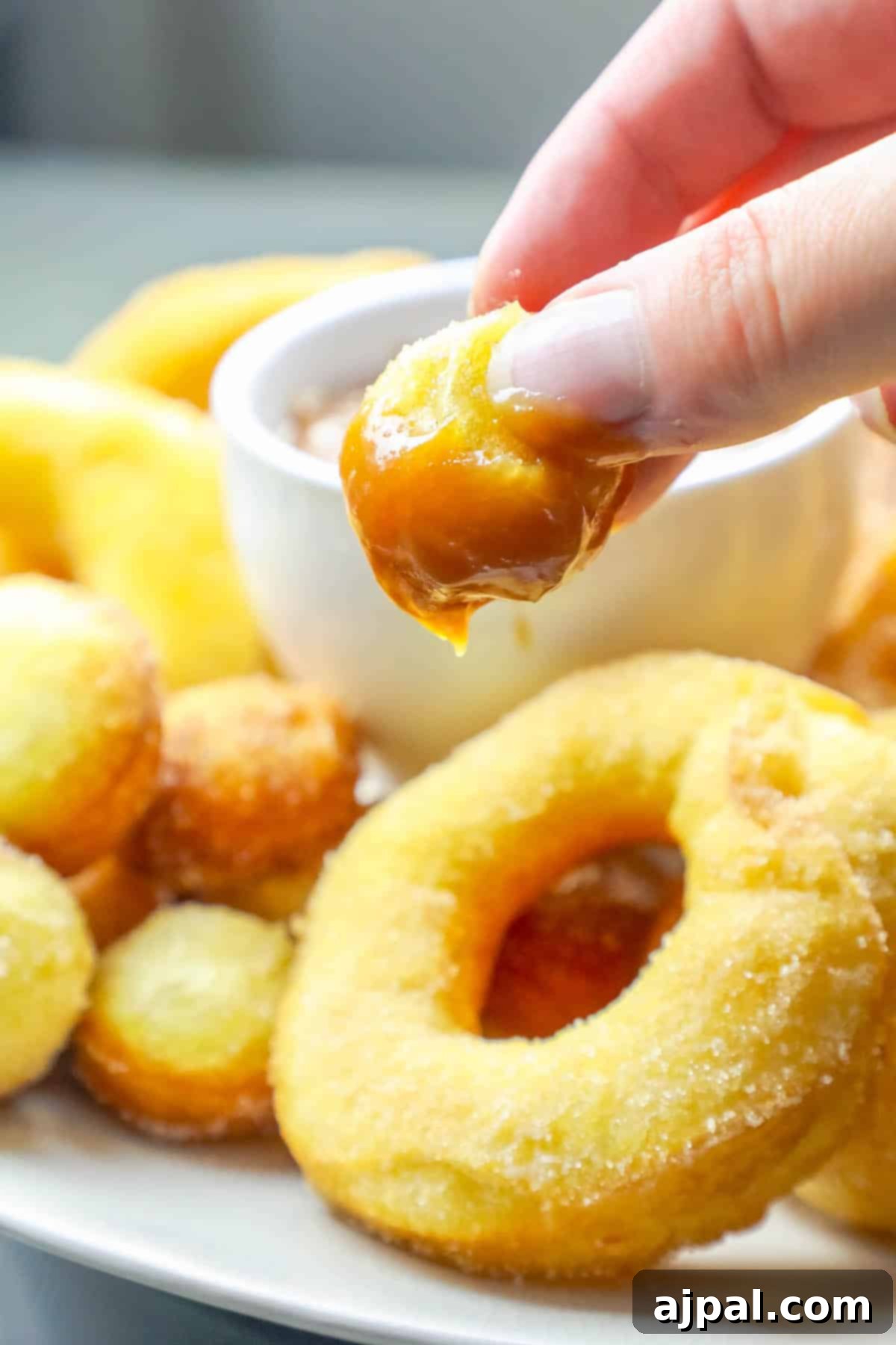 A hand gently dipping a freshly fried, mulling spiced donut into a creamy, golden caramel sauce. The focus is on the perfect texture of the donut and the rich sauce.