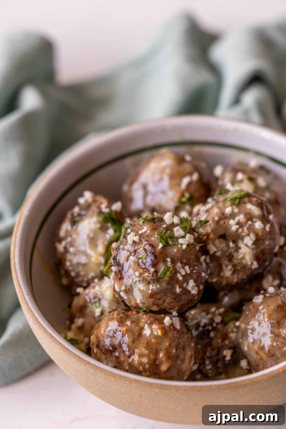 A beautifully presented bowl of Garlic Butter Parmesan Meatballs, garnished with grated cheese and fresh chopped herbs, resting on a light surface with a soft green cloth in the background.