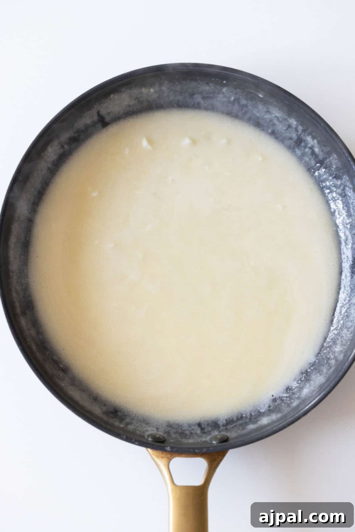 A frying pan on a stovetop contains a thick white sauce, simmering over low heat, forming a delicious foundation for the meatballs.