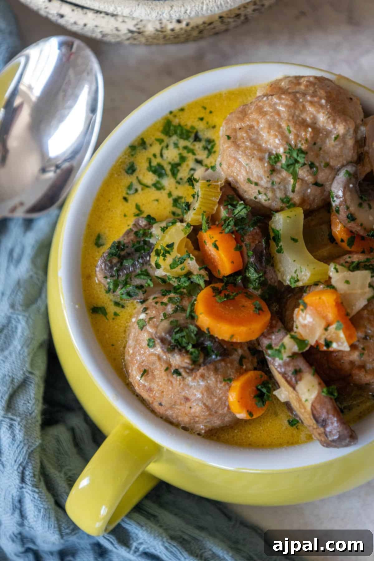 Rich & Creamy One-Pot Meatball Delight 13 An easy meatball soup recipe served in a yellow bowl, featuring savory meatballs, sliced carrots, celery, and mushrooms garnished with herbs. A spoon rests nearby on a blue cloth.