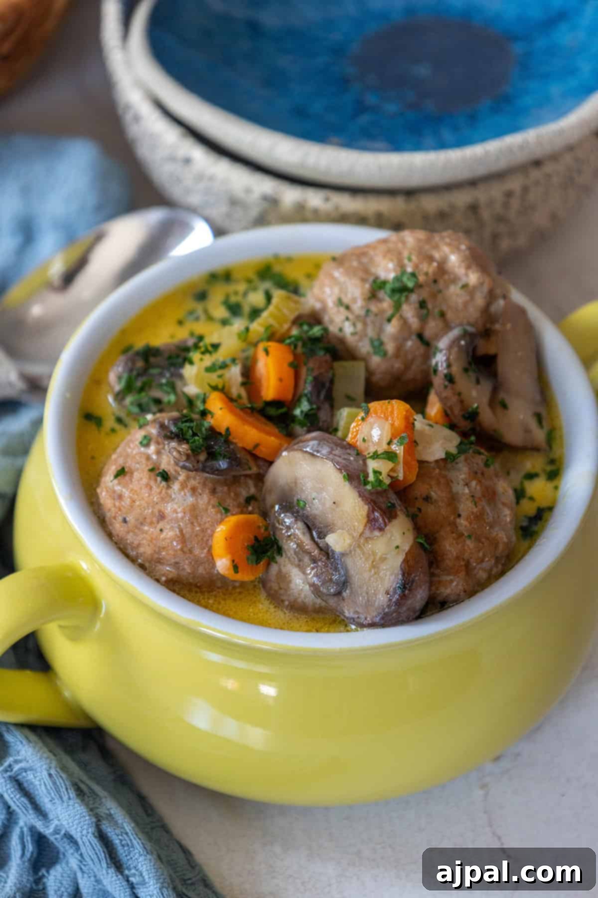 Rich & Creamy One-Pot Meatball Delight 3 A yellow bowl brimming with a one pot meatball soup recipe, featuring tender meatballs, mushrooms, carrots, and herbs in a creamy sauce. It sits invitingly on the table beside a blue towel and ceramic bowls.