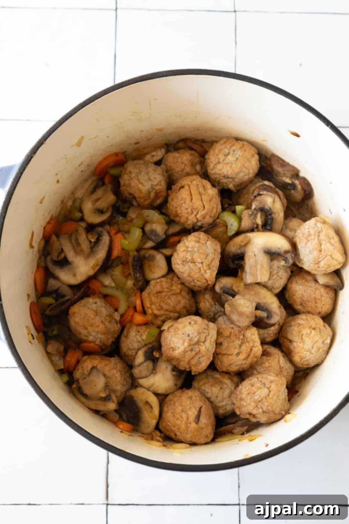 Rich & Creamy One-Pot Meatball Delight 8 A pot filled with meatballs, sliced mushrooms, carrots, and celery sits on a white tiled surface—a perfect start to an easy meatball recipe that transforms into a creamy one-pot meatball soup.
