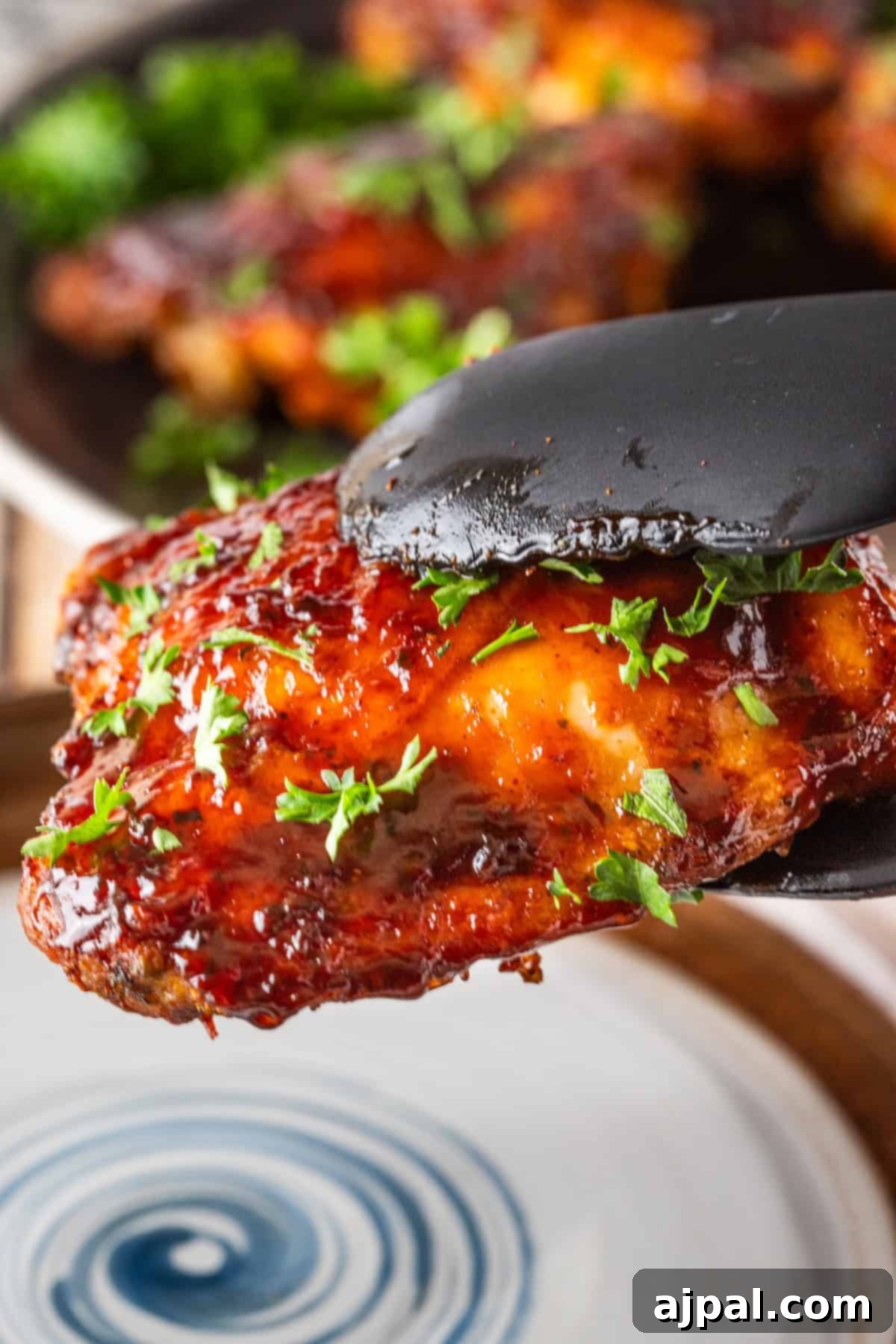 Sizzling Skillet BBQ Chicken Thighs 2 Close-up of black tongs holding a glazed, grilled chicken wing garnished with chopped parsley. Other wings are blurred in the background on a dark plate, reminiscent of those sizzling keto BBQ chicken thighs pan-fried to perfection.