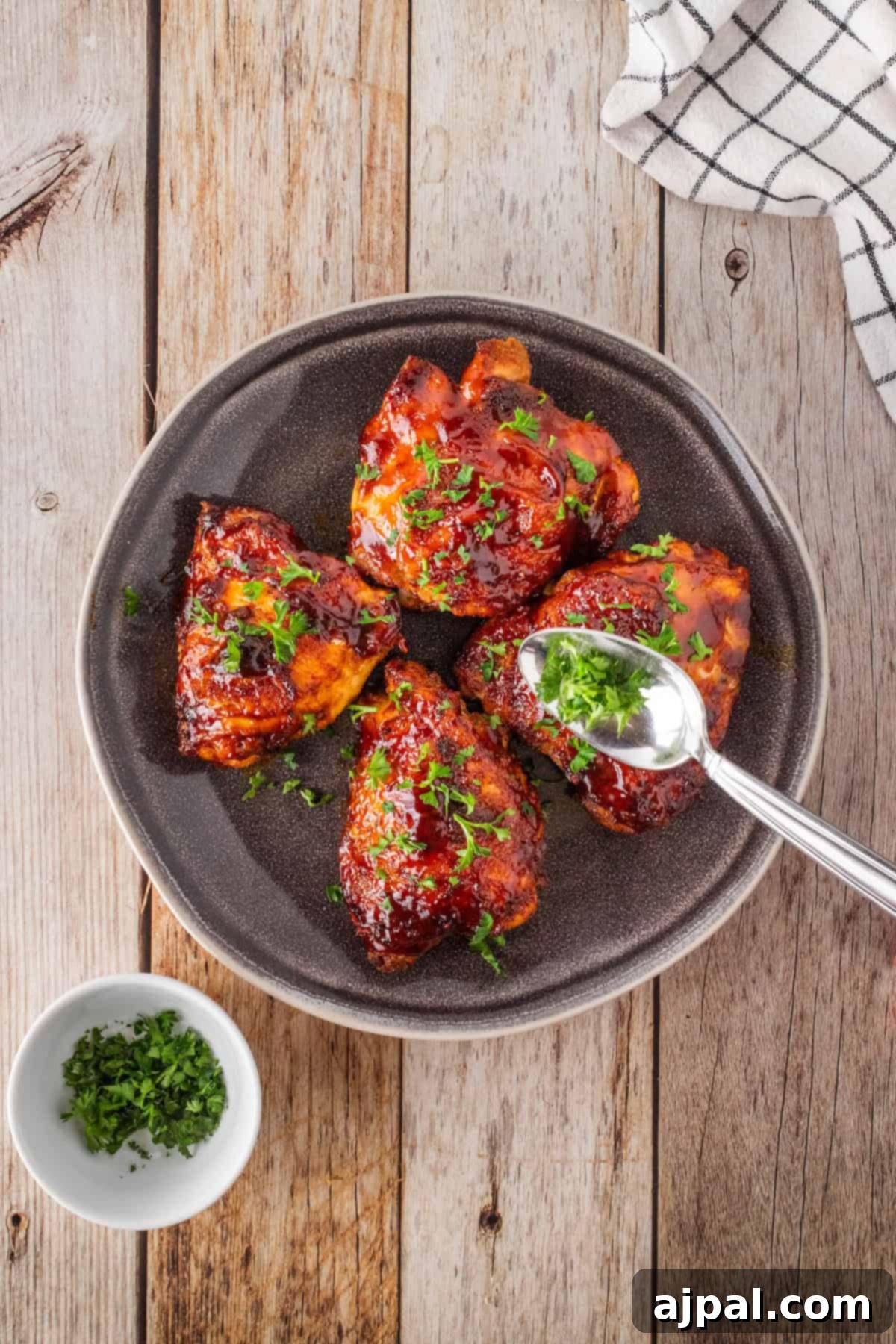 Sizzling Skillet BBQ Chicken Thighs 7 A plate of keto BBQ chicken thighs gleams with glaze and a sprinkle of parsley. A spoon offers extra herbs, poised beside a small bowl of parsley on the wooden table. Easy one pan BBQ chicken thighs and a checkered cloth complete this inviting scene.