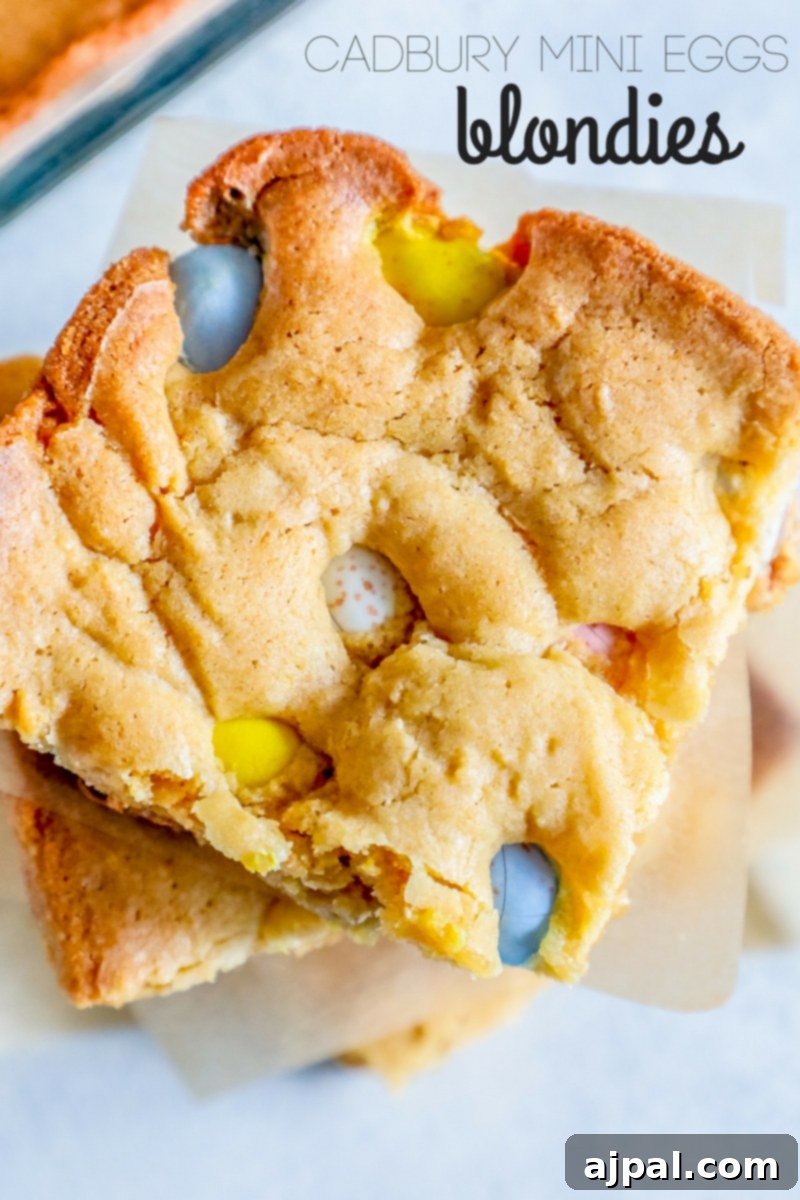 Gooey blondie bars studded with vibrant Cadbury Mini Eggs, stacked on a white plate, highlighting their rich texture and colorful candy inclusions.