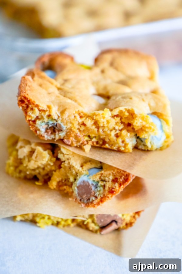 A stack of golden blondie bars, generously studded with colorful Cadbury Mini Eggs and chocolate chips, resting on a rustic wooden surface, ready to be enjoyed.